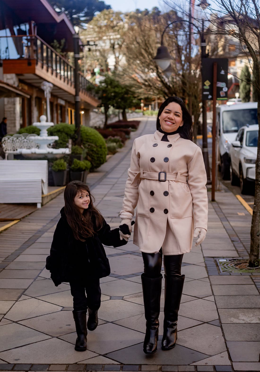 Mãe e filha de mãos dadas, sorrindo, numa rua arborizada
